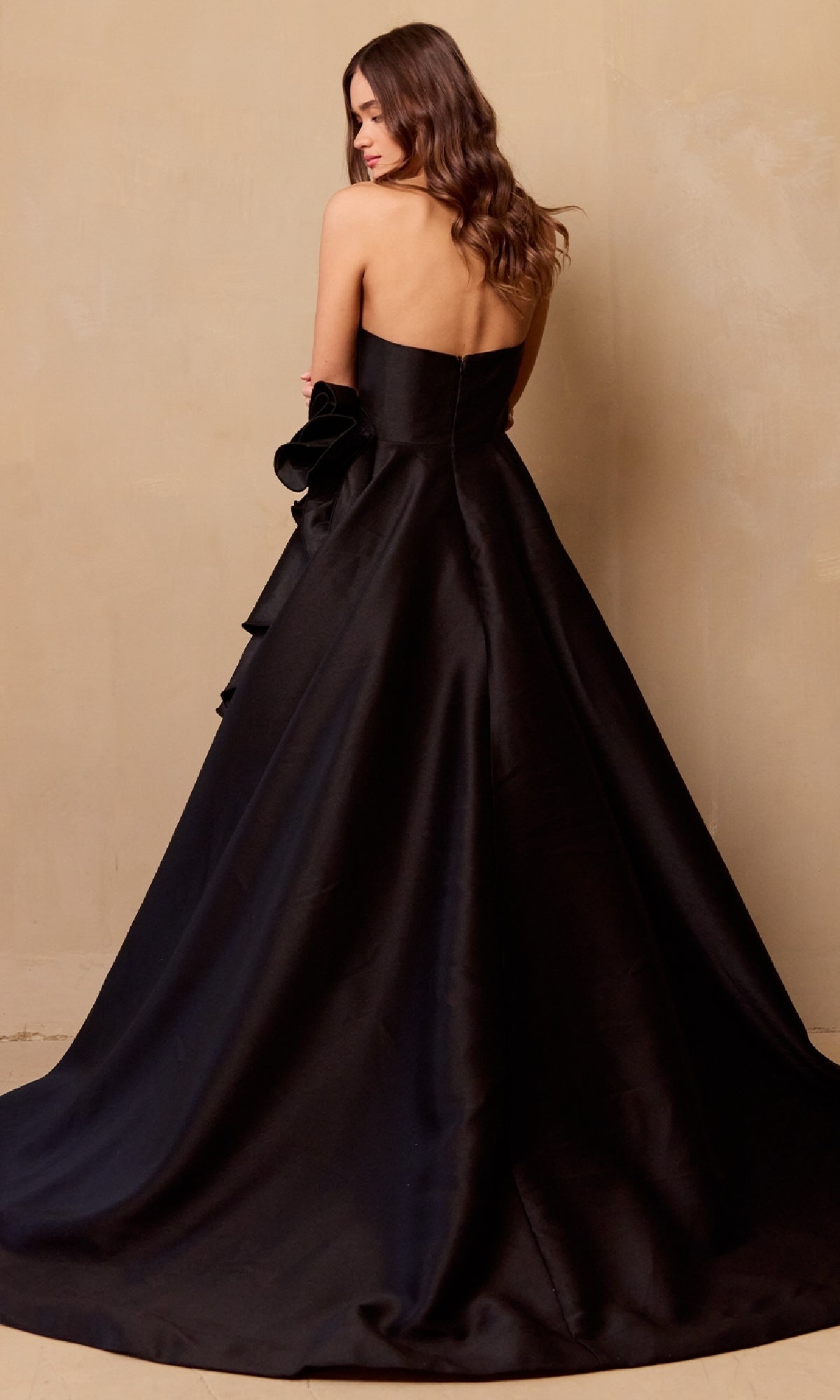 A woman stands with her back to the camera against a plain beige wall, wearing the Amelia Couture SU085 Strapless Prom Ball Gown: a floor-length black sweetheart dress with a flared skirt.